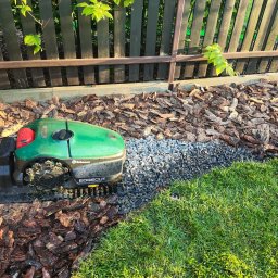 ProfGrass - Robot koszący Robomow EdgeCut na granicy trawnika, kory i drenażu żwirowego przy drewnianym płocie.