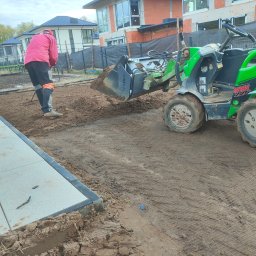 ProfGrass - Przygotowanie podłoża pod kostkę brukową: miniładowarka rozrzuca ziemię, a pracownik wyrównuje ją grabiami na siatce wzmacniającej.