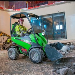 ProfGrass - Operator w kasku i kamizelce odblaskowej obsługuje zieloną mini-ładowarkę Avant 225, zbierając gruz wewnątrz okrągłego budynku w trakcie prac rozbiórkowych.