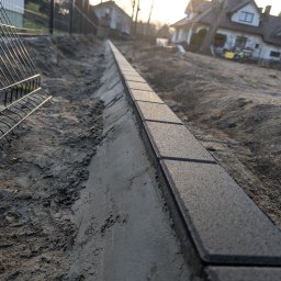 ProfGrass - Krawężnik z kostki brukowej na świeżym betonie, widok z dołu, w tle dom i ogrodzenie. Ułożony brzeg chodnika w trakcie prac ziemnych. Słoneczny dzień.