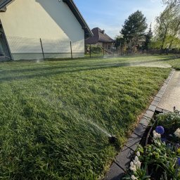 ProfGrass - Automatyczne nawadnianie trawnika w ogrodzie, zraszacz w akcji. W tle dom jednorodzinny i ogrodzenie. Widoczne kwiaty wzdłuż ścieżki.