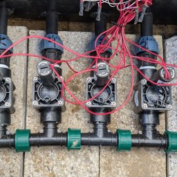 ProfGrass - System nawadniania: cztery zawory Hunter PGV z przewodami elektrycznymi, zainstalowane na zewnątrz, na betonowych płytach. Widoczne oznaczenia numerów zaworów.