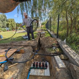 ProfGrass - Instalacja systemu nawadniania w ogrodzie: pracownik układa rury w wykopie, widoczne złącza i narzędzia. W tle zielona trawa i drzewa.