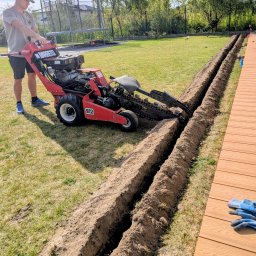 ProfGrass - Czerwona glebogryzarka 'Barreto 912' tworzy równoległe rowy w trawniku obok drewnianego tarasu. Operator w czapce nadzoruje pracę. W tle dom i trampolina.