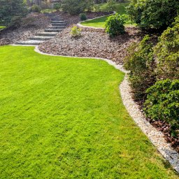 ProfGrass - Zadbany ogród z intensywnie zielonym trawnikiem, ścieżką z kamiennych stopni i rabatami wysypanymi korą, obrzeża z białych kamyczków, krzewy. Słoneczny dzień.