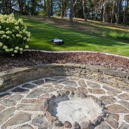 ProfGrass - Ogród z paleniskiem wyłożonym kamieniami i białym żwirem, obrzeża z kory drzewnej i kamienny murek. W tle trawnik z robotem koszącym i drzewa.