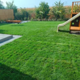 Świeżo założony trawnik z rolki, widok na plac zabaw z drewnianą zjeżdżalnią, obrzeża z kostki brukowej i drewnianą piaskownicę. Słoneczny dzień, zadbany ogród.