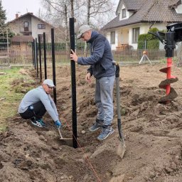 ProfGrass - Montaż ogrodzenia: Dwóch mężczyzn ustawia słupki w ziemi, używając poziomicy i szpadla. W tle widoczna żółta wiertnica do ziemi i zabudowa jednorodzinna.