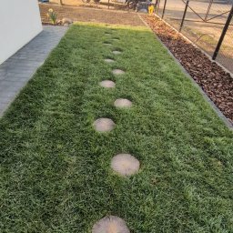 ProfGrass - Ścieżka z drewnianych krążków przez trawnik, obok domu z kostką brukową i korą, w tle samochód dostawczy i toaleta przenośna.