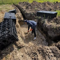 ProfGrass - Wykop w ziemi z drenażem żwirowym, przygotowany pod montaż skrzynek rozsączających. Pracownik wyrównuje podłoże. W tle zabudowa jednorodzinna.