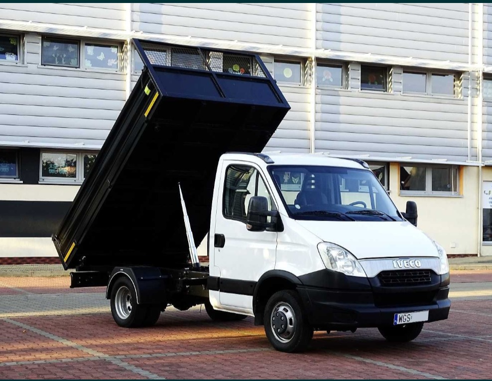 Biała wywrotka Iveco z podniesioną czarną skrzynią ładunkową stoi na czerwonej kostce brukowej przed budynkiem z szarej blachy falistej.