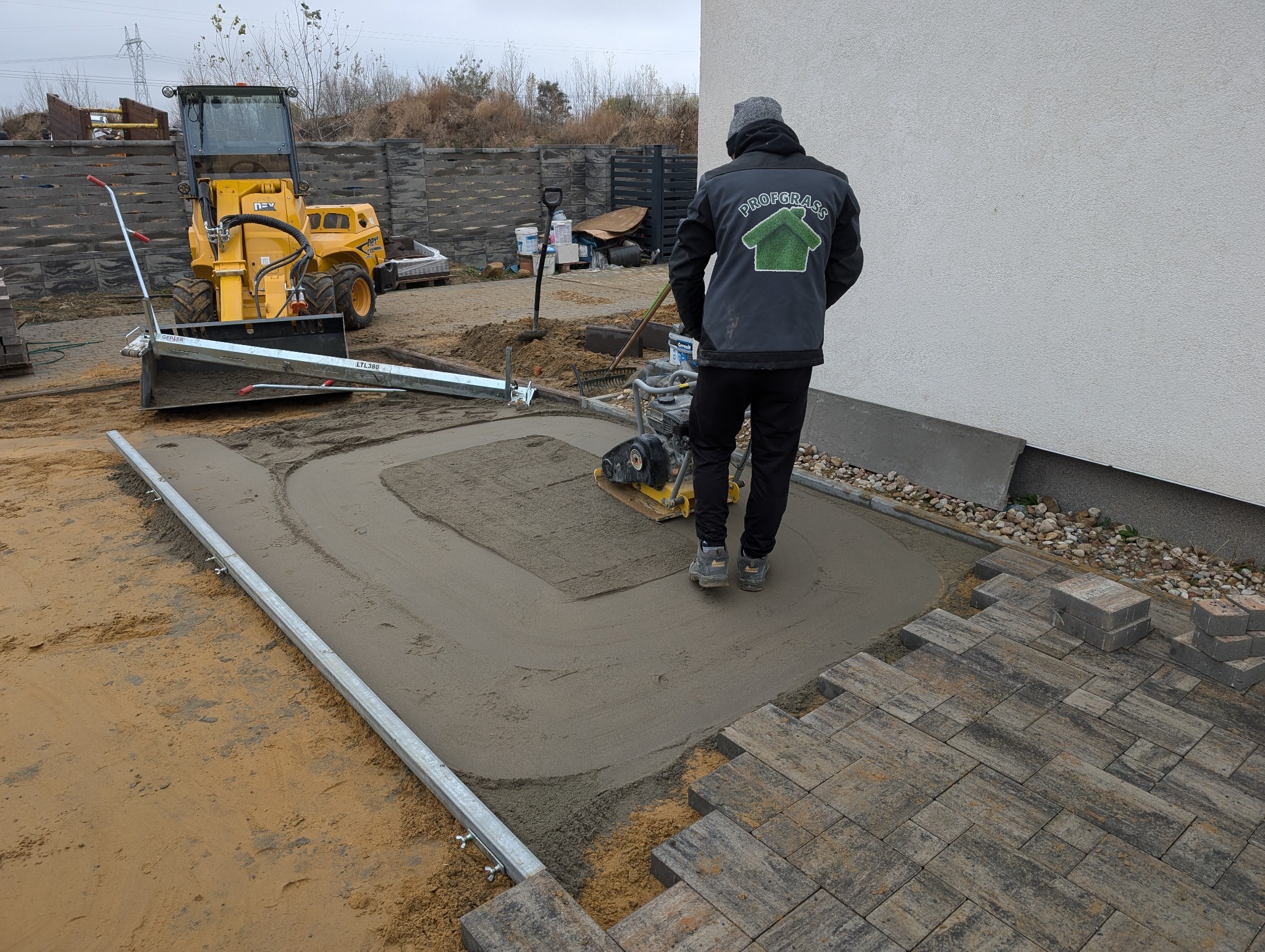 Pracownik w kurtce z logo firmy PROFGASS zagęszcza piasek pod kostkę brukową za pomocą zagęszczarki spalinowej; w tle żółta miniładowarka i przygotowany teren.