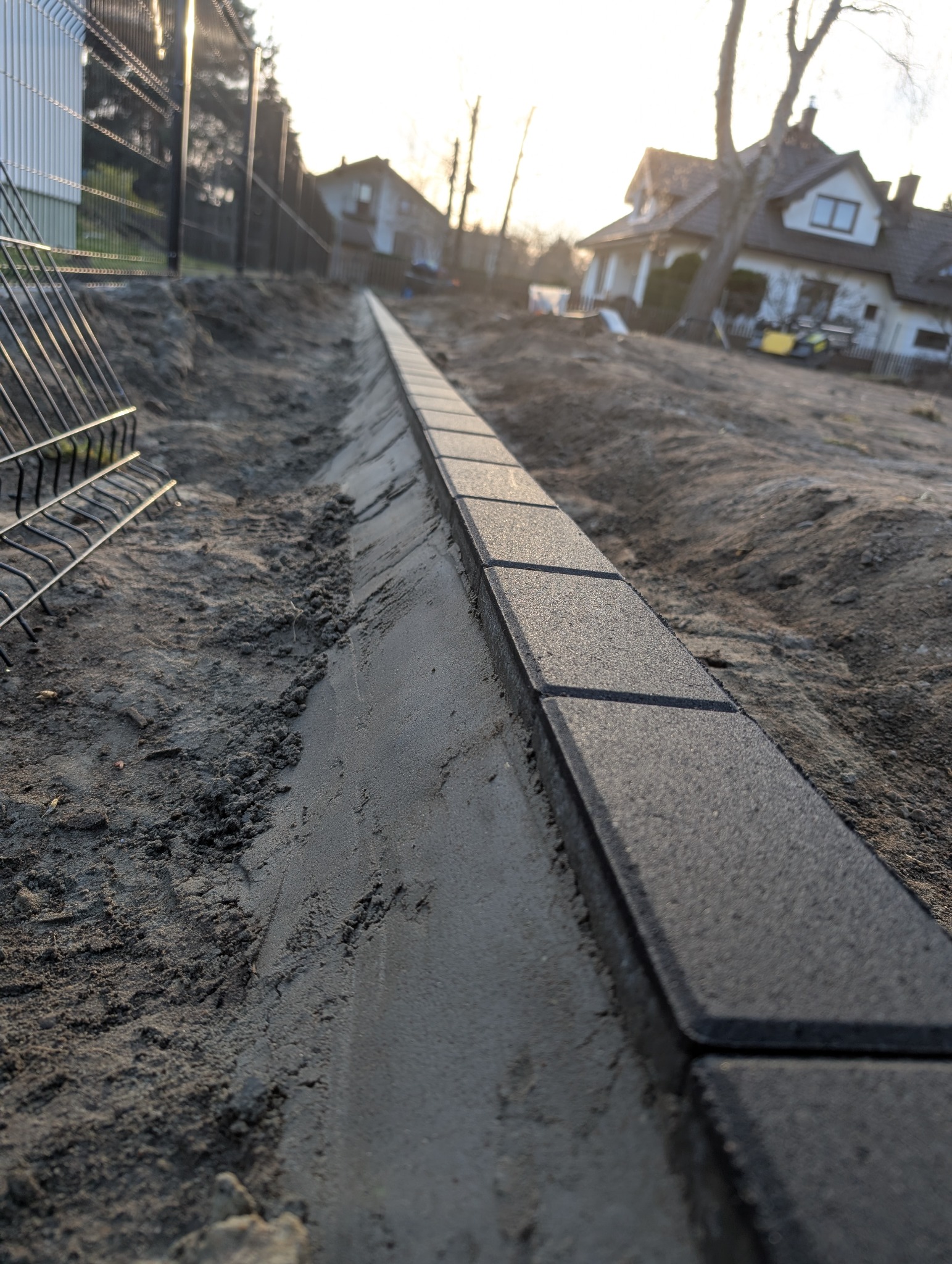Krawężnik z kostki brukowej na świeżym betonie, widok z dołu, w tle dom i ogrodzenie. Ułożony brzeg chodnika w trakcie prac ziemnych. Słoneczny dzień.
