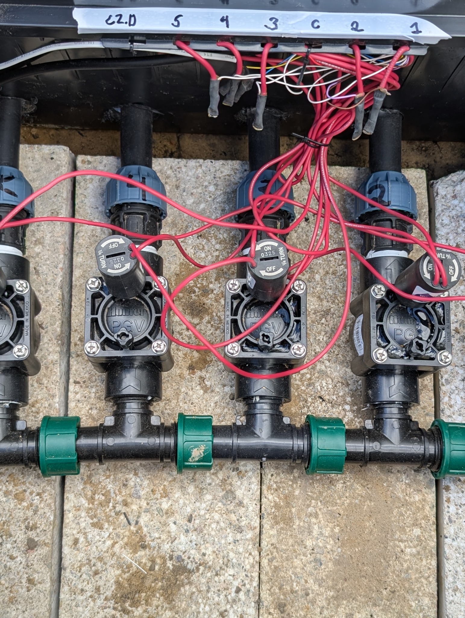 System nawadniania: cztery zawory Hunter PGV z przewodami elektrycznymi, zainstalowane na zewnątrz, na betonowych płytach. Widoczne oznaczenia numerów zaworów.