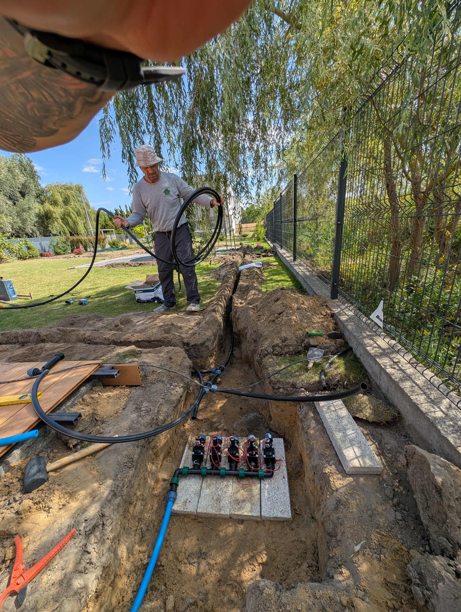 Instalacja systemu nawadniania w ogrodzie: pracownik układa rury w wykopie, widoczne złącza i narzędzia. W tle zielona trawa i drzewa.