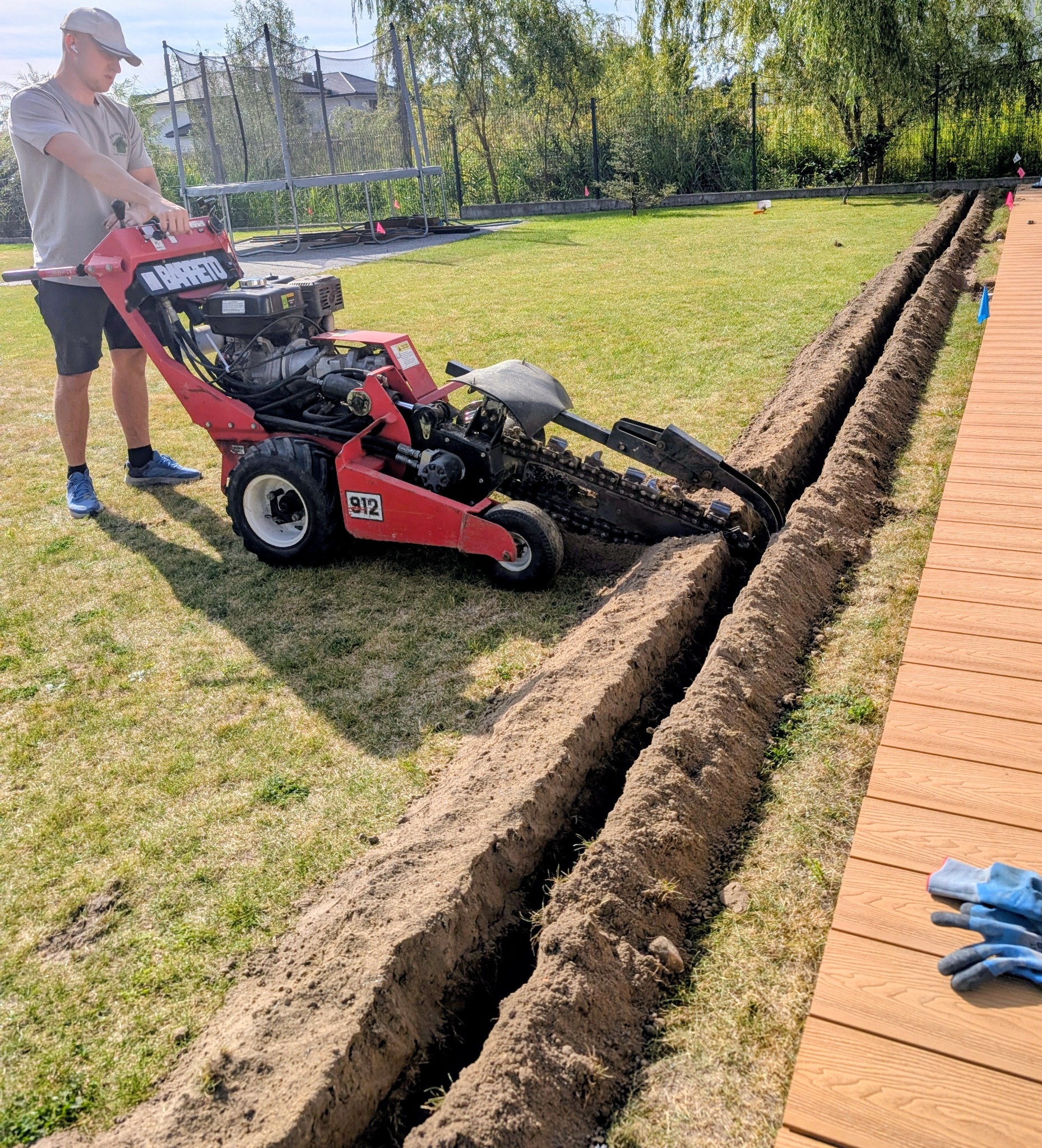 Czerwona glebogryzarka 'Barreto 912' tworzy równoległe rowy w trawniku obok drewnianego tarasu. Operator w czapce nadzoruje pracę. W tle dom i trampolina.