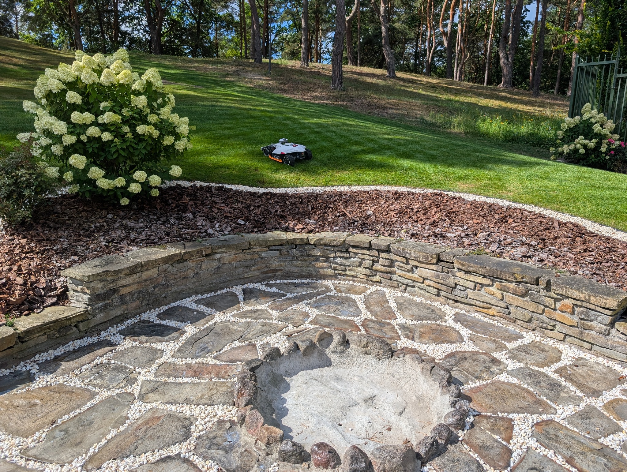 Ogród z paleniskiem wyłożonym kamieniami i białym żwirem, obrzeża z kory drzewnej i kamienny murek. W tle trawnik z robotem koszącym i drzewa.