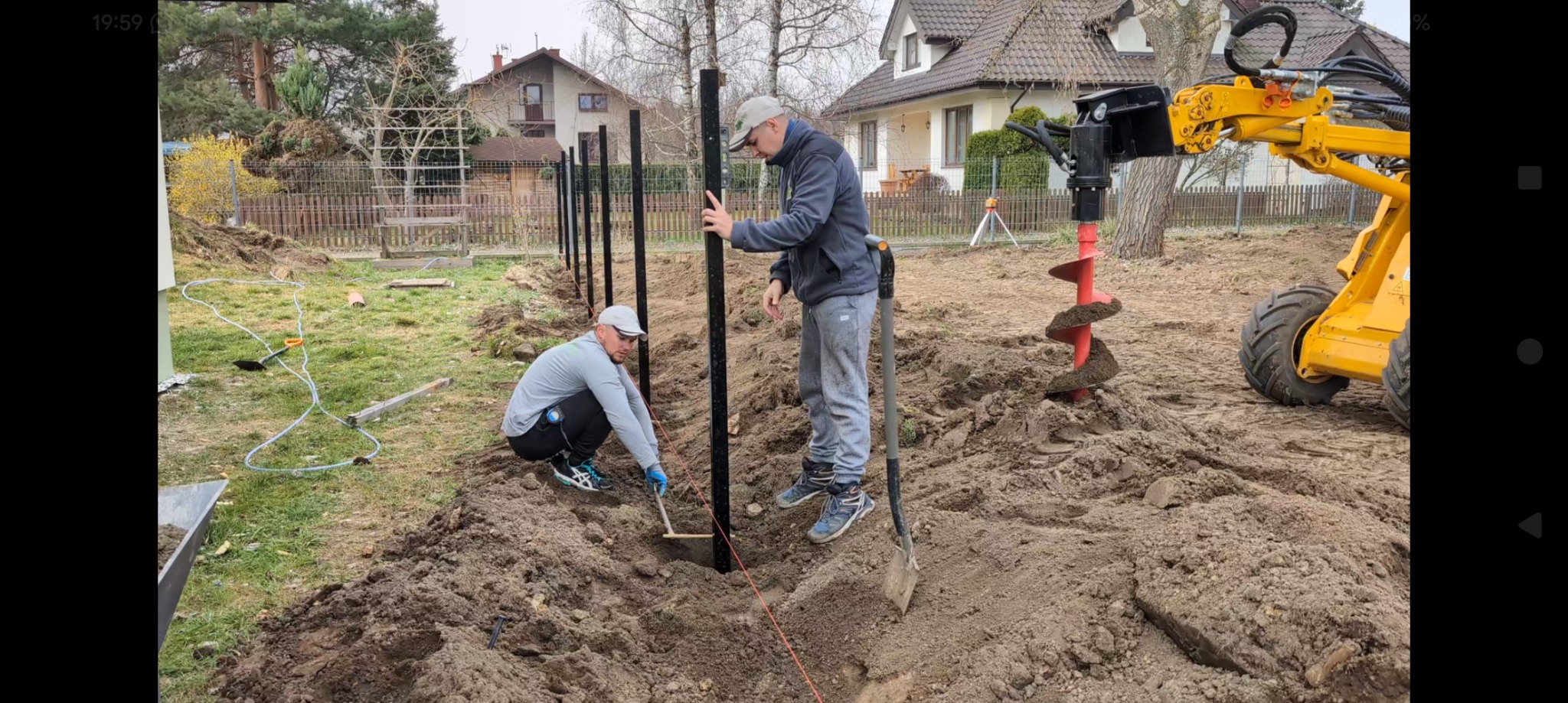 Montaż ogrodzenia: Dwóch mężczyzn ustawia słupki w ziemi, używając poziomicy i szpadla. W tle widoczna żółta wiertnica do ziemi i zabudowa jednorodzinna.