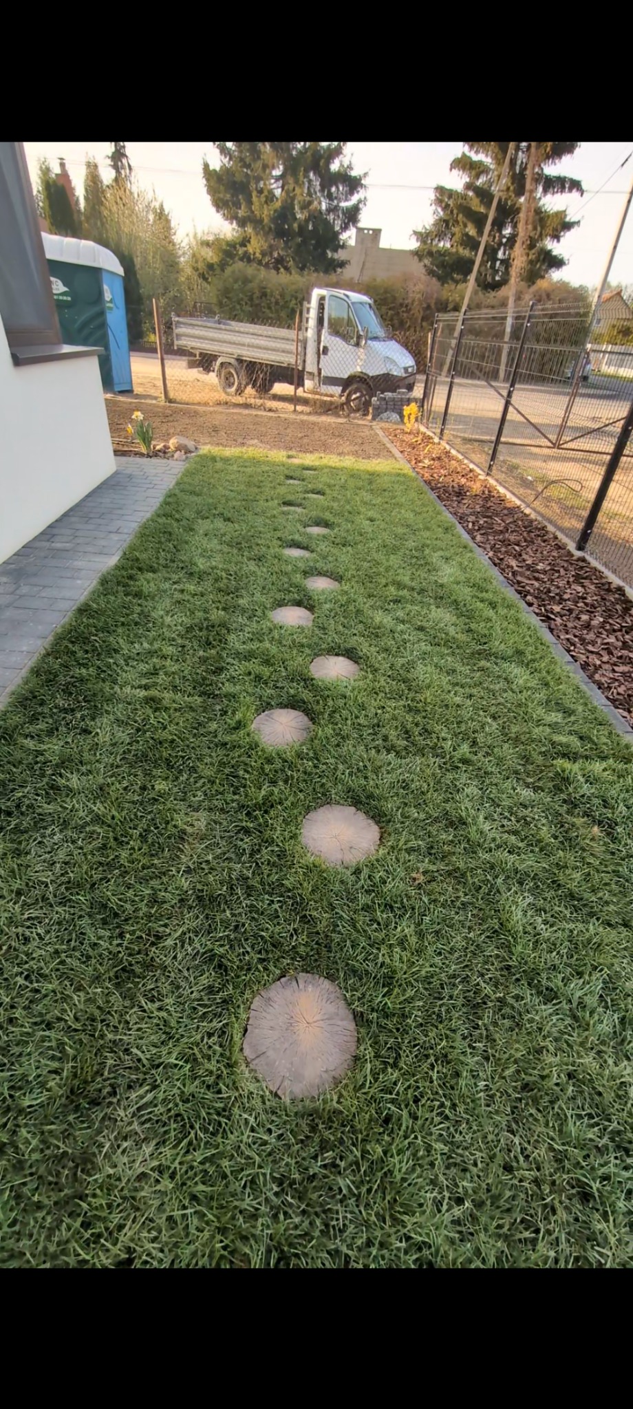 Ścieżka z drewnianych krążków przez trawnik, obok domu z kostką brukową i korą, w tle samochód dostawczy i toaleta przenośna.