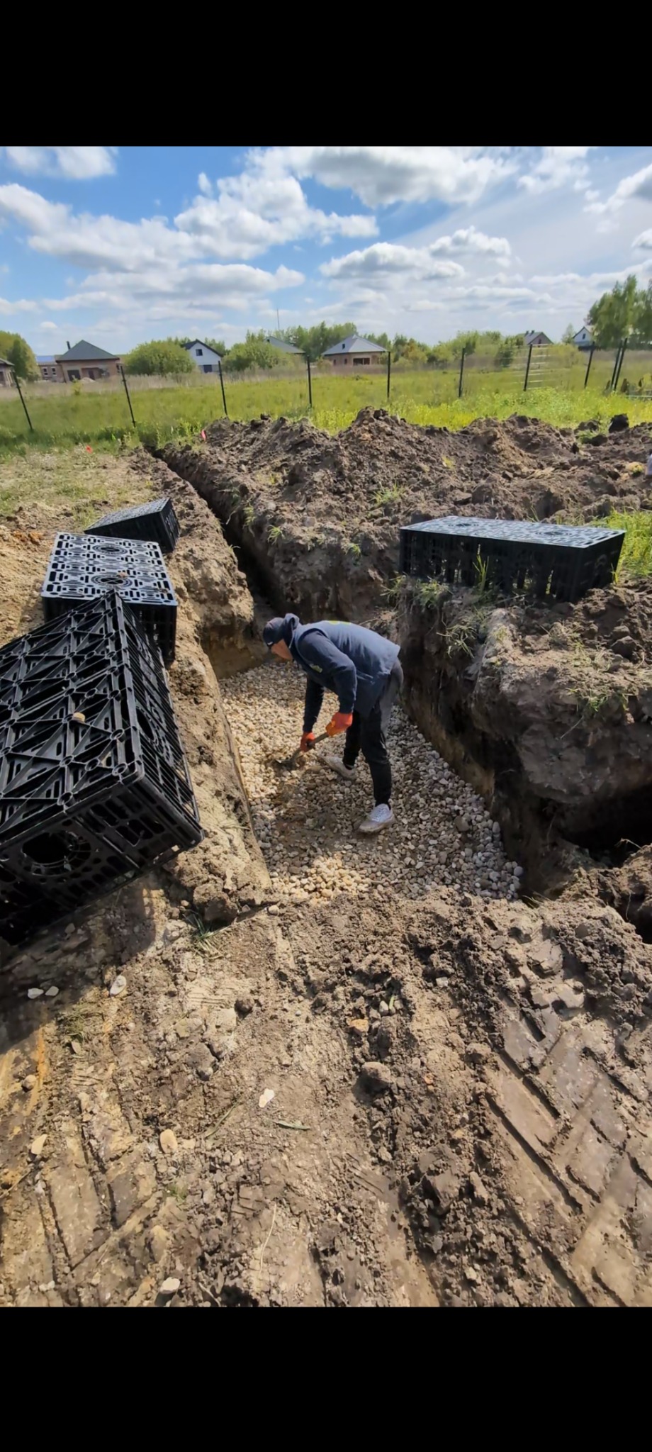 Wykop w ziemi z drenażem żwirowym, przygotowany pod montaż skrzynek rozsączających. Pracownik wyrównuje podłoże. W tle zabudowa jednorodzinna.