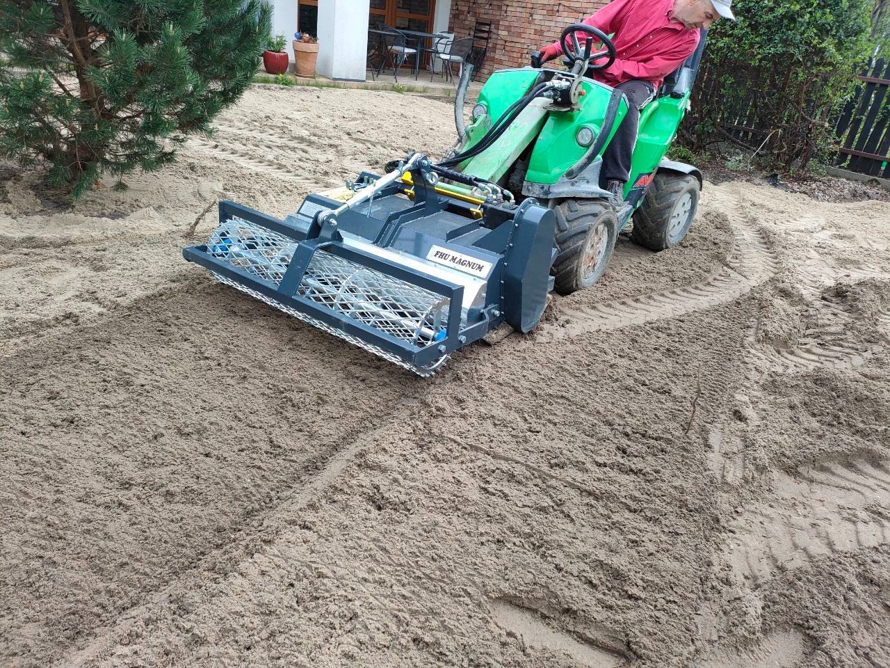 Operator wyrównuje teren piaskowy specjalistycznym traktorem z walcem siatkowym, przygotowując podłoże pod trawnik w ogrodzie.