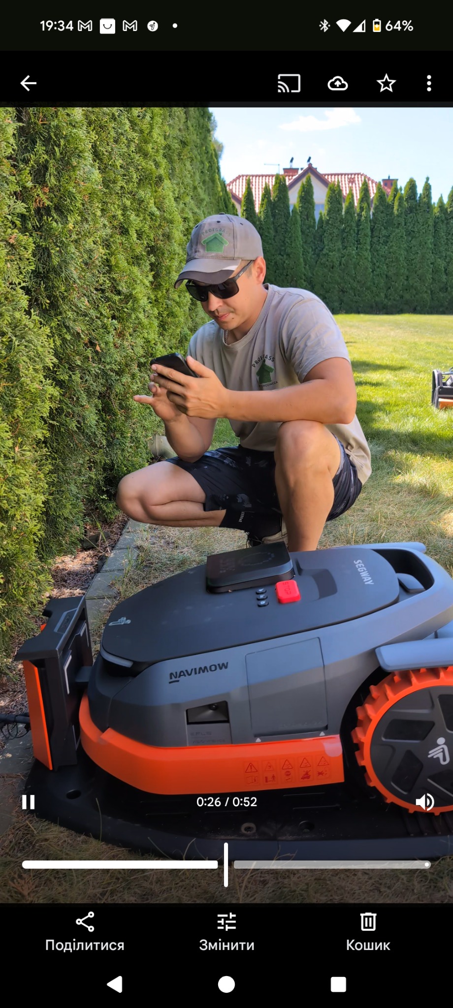Mężczyzna w czapce z logo firmy ogrodniczej, kuca obok automatycznej kosiarki Segway Navimow, patrząc na telefon. W tle żywopłot z tuj.