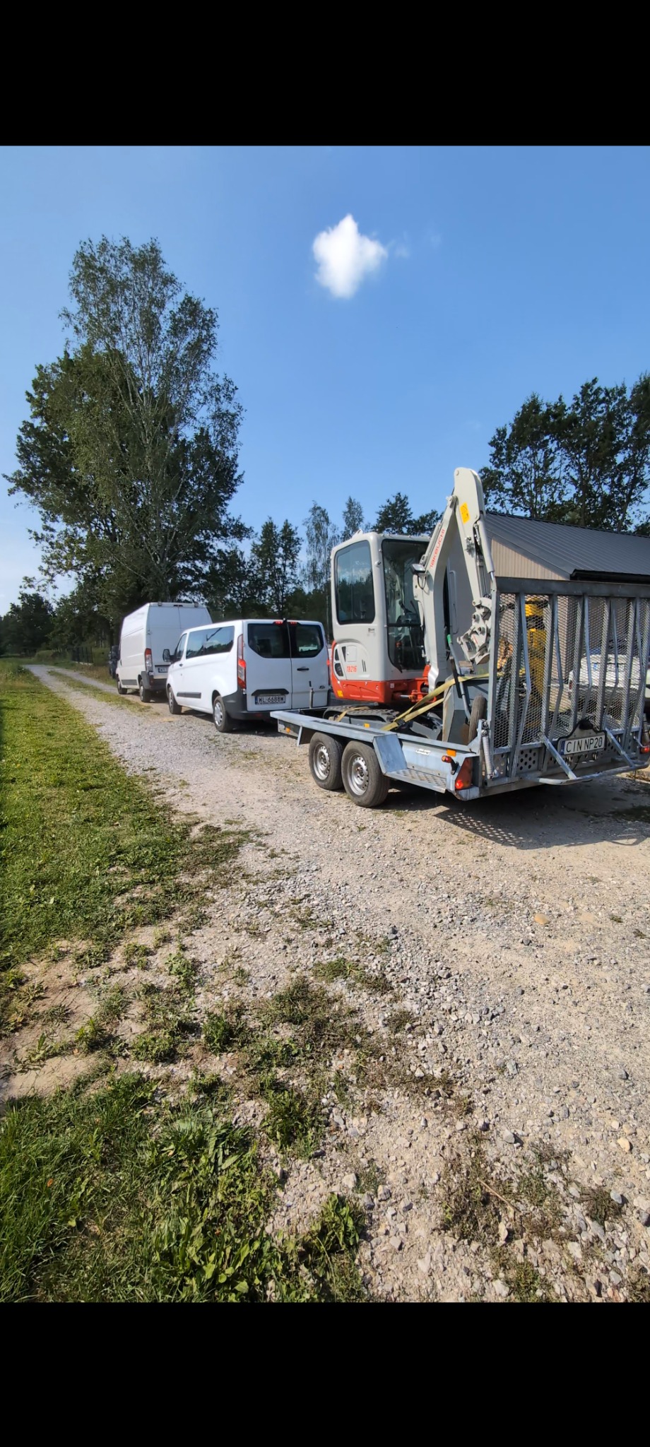 Pomarańczowa minikoparka TEREX na przyczepie, zaparkowana obok trzech białych busów na żwirowej drodze w słoneczny dzień. Widoczne drzewa i błękitne niebo.