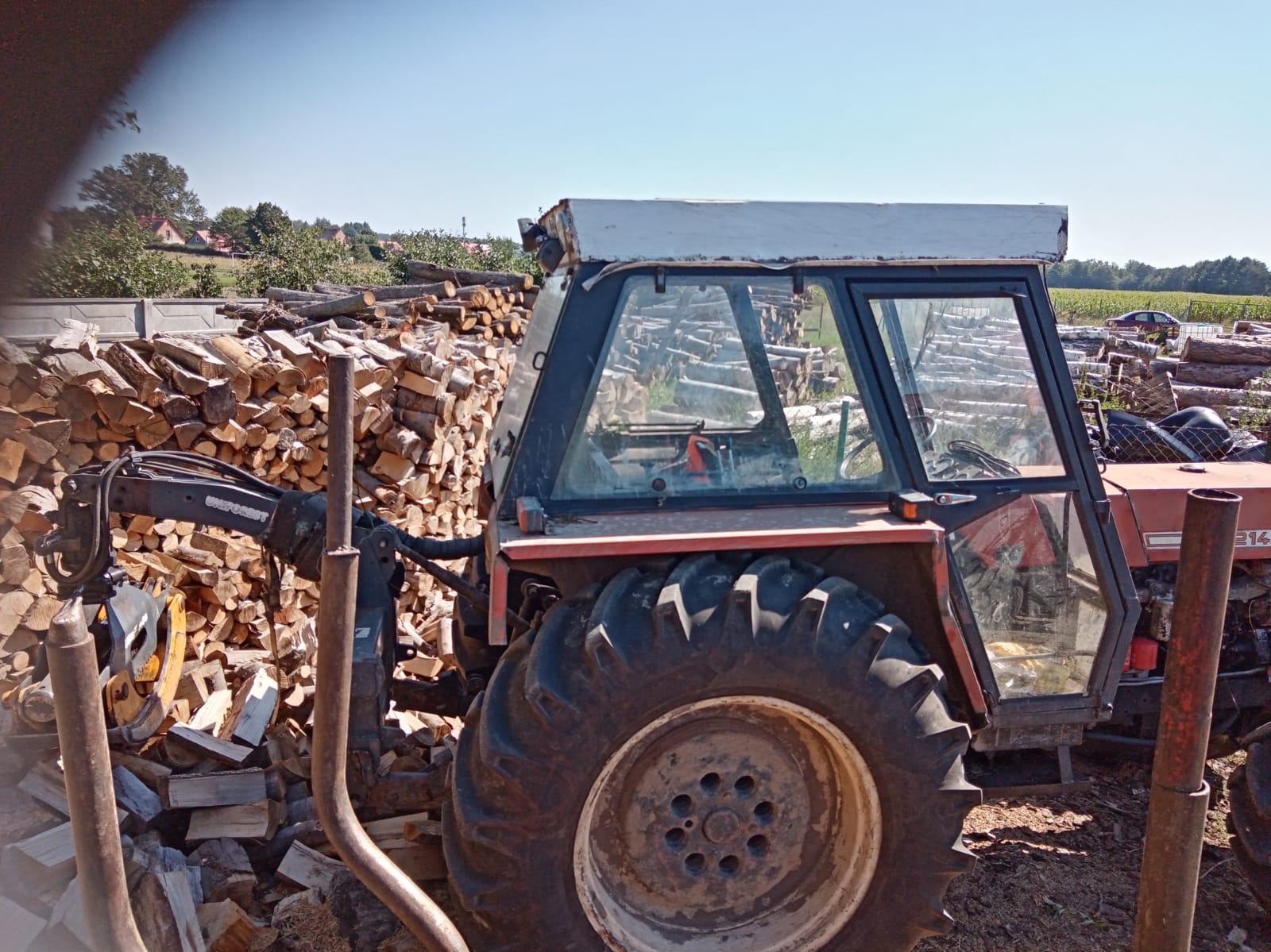 Czerwony traktor z chwytakiem hydraulicznym stoi obok stosu pociętego drewna na tle zieleni i błękitnego nieba.