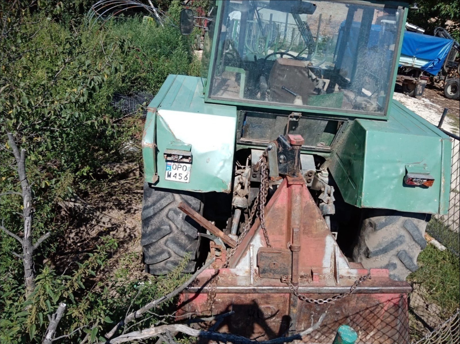 Zielony traktor z zamontowanym urządzeniem do karczowania pni, otoczony zielenią i gałęziami, widok z góry.