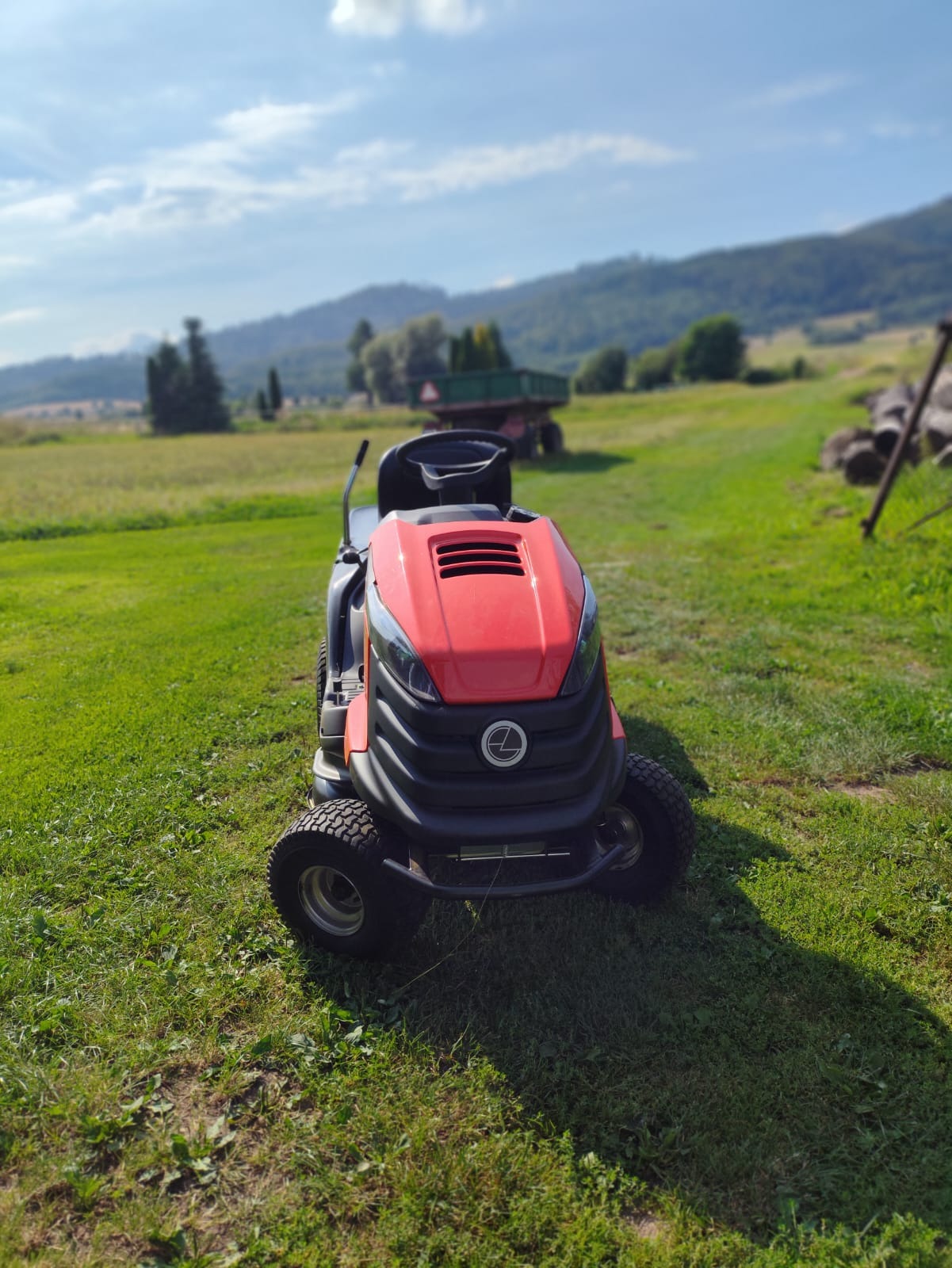 Czerwono-czarny traktorek do trawy stoi na zielonym trawniku, w tle pole, góry i przyczepa.