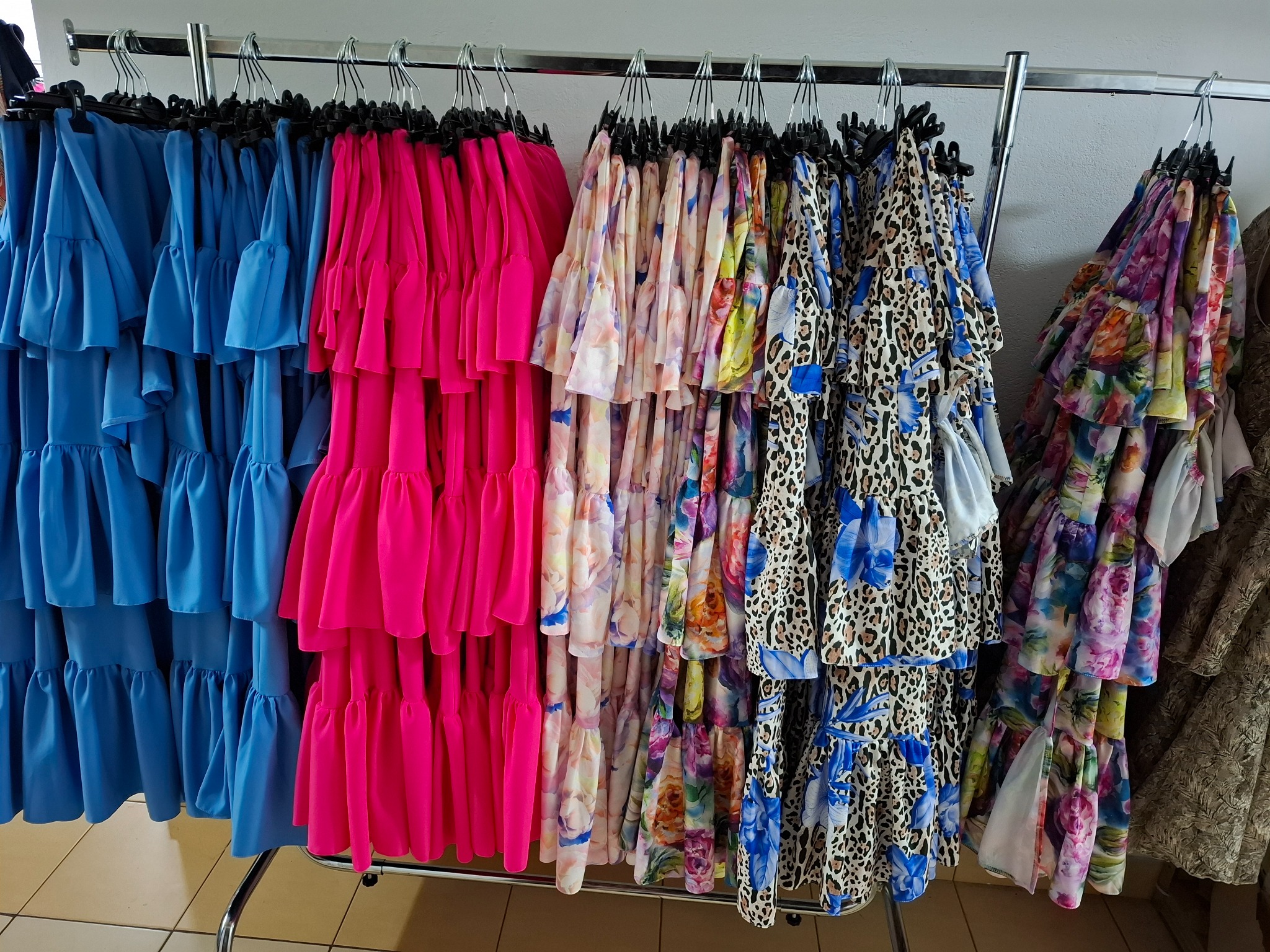 Różnokolorowe, warstwowe sukienki z falbanami wiszące na metalowym wieszaku w szwalni, widoczne odcienie niebieskiego, różu, kwiatowe wzory i panterka.