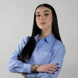 Portret kobiety w błękitnej koszuli w paski z założonymi rękami i złotym zegarkiem na tle jasnej ściany.