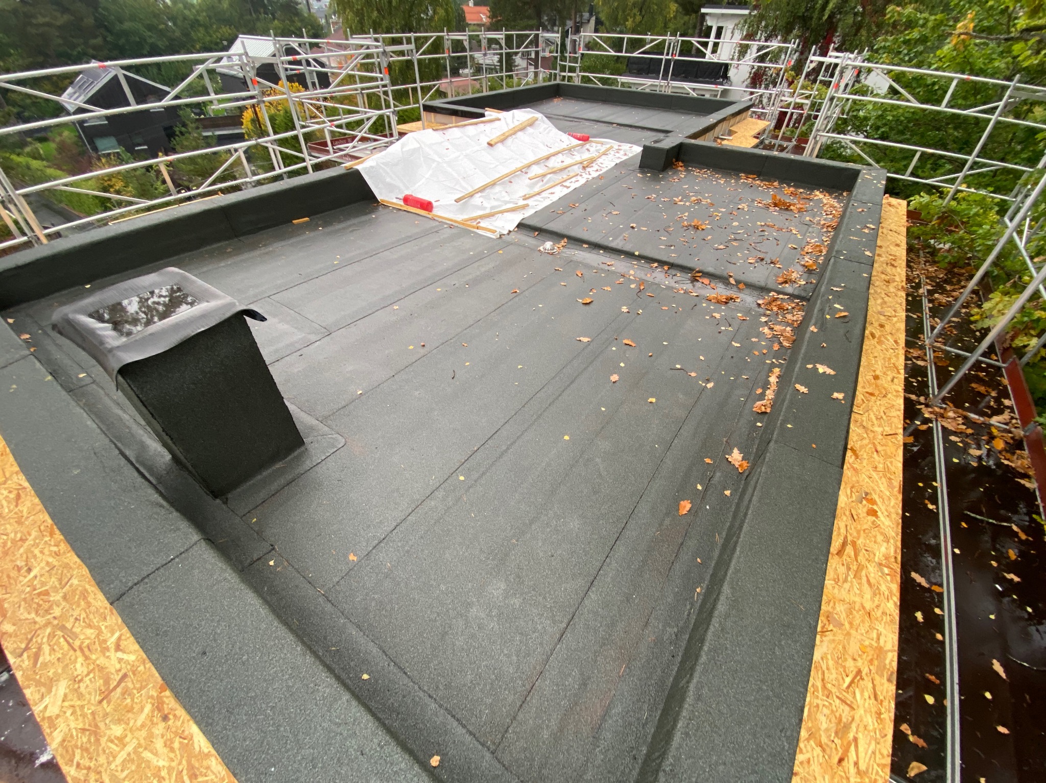Płaski dach pokryty papą, częściowo zabezpieczony folią i drewnianymi belkami, z kominem i elementami rusztowania. Widoczne jesienne liście.