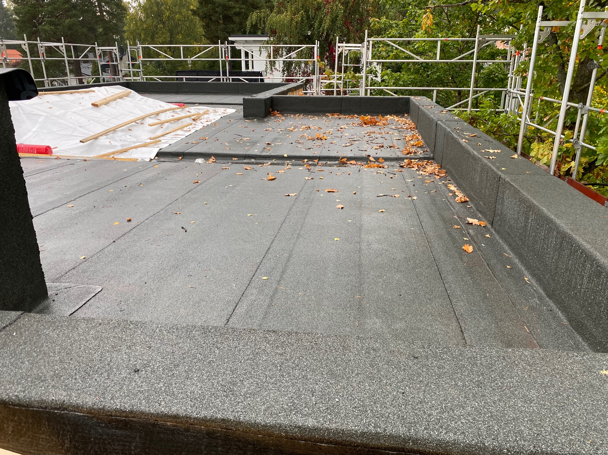 Płaski dach pokryty papą, z widocznymi obróbkami blacharskimi, częściowo zakryty folią i drewnianymi belkami, na tle rusztowania i zieleni.