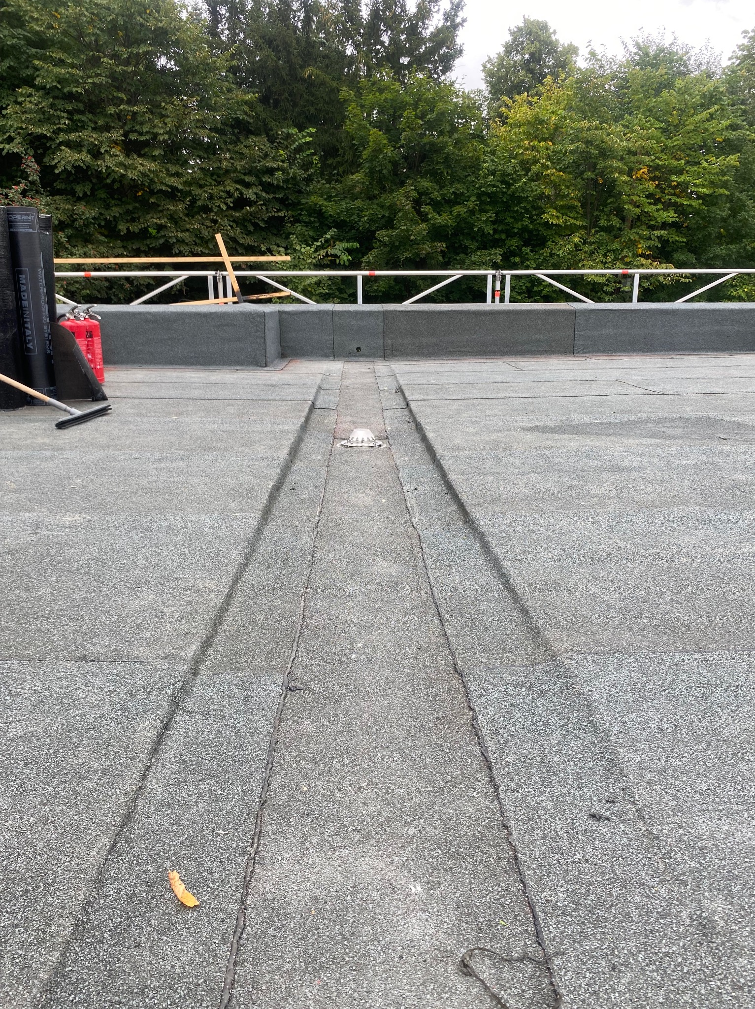 Płaski dach pokryty papą z odpływem liniowym. Widoczne rolki papy, drabina i narzędzia. Tło: drzewa.