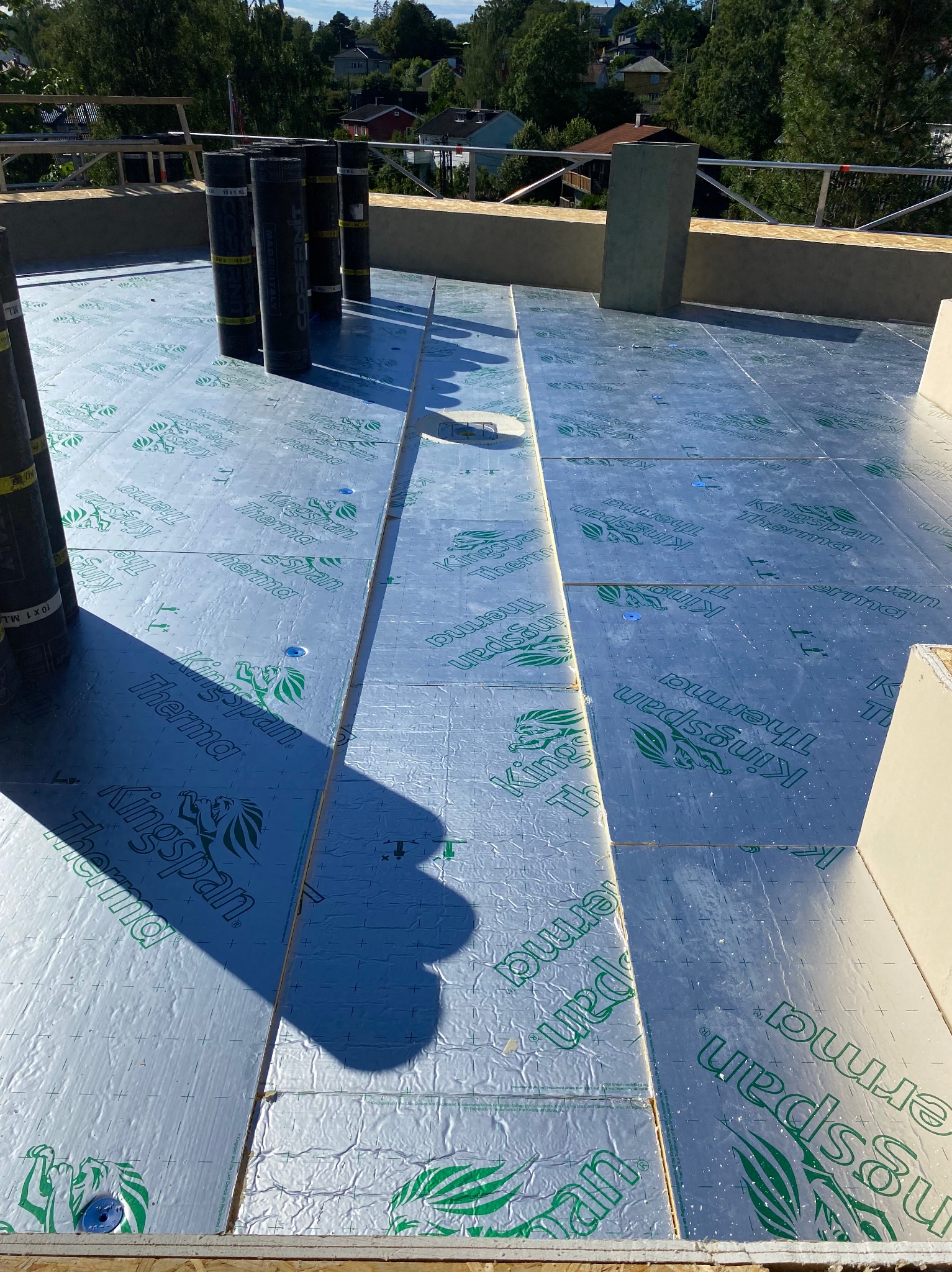 Płyty izolacyjne Kingspan Therma na dachu w trakcie montażu. Widoczne rolki papy i elementy konstrukcji. Jasne światło słoneczne.