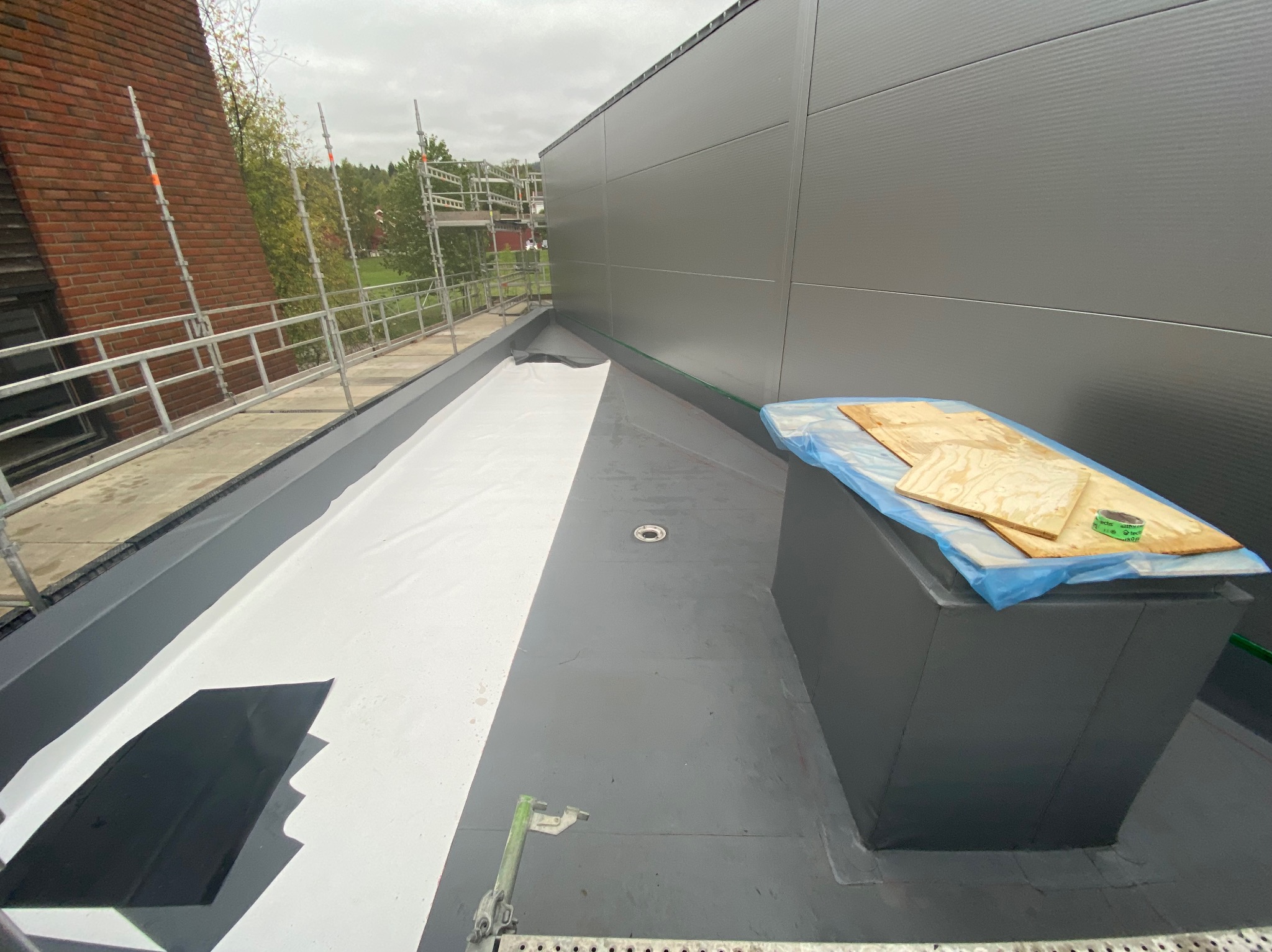 Wodoodporna izolacja dachu płaskiego z detalami wykończeniowymi, widok z góry. Na dachu leżą deski i narzędzia, a w tle rusztowanie i budynek.