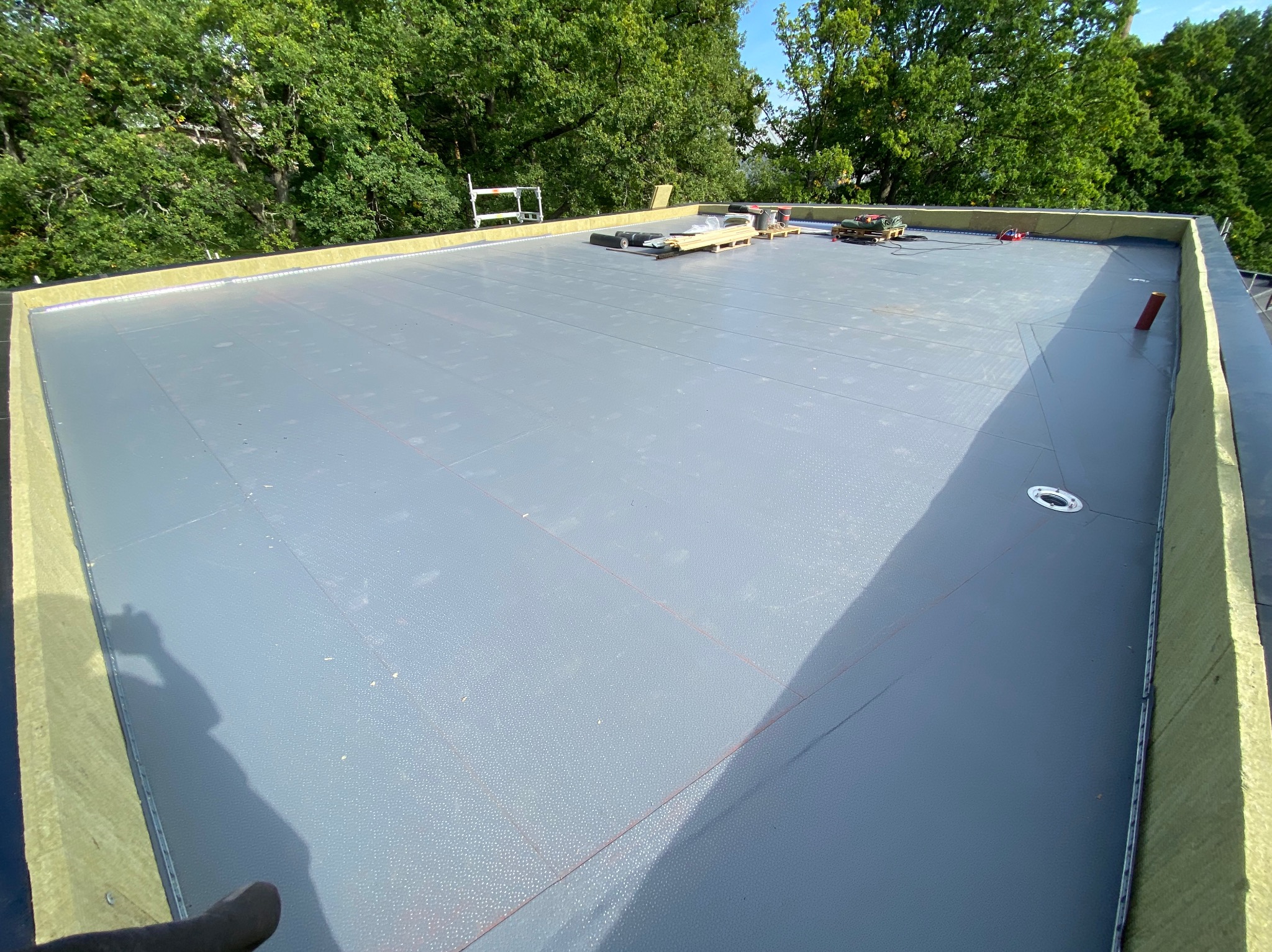 Płaski dach pokryty szarą membraną dachową, z widoczną izolacją termiczną na krawędziach. Na dachu narzędzia, palety i drabina. Tło: zielone drzewa.