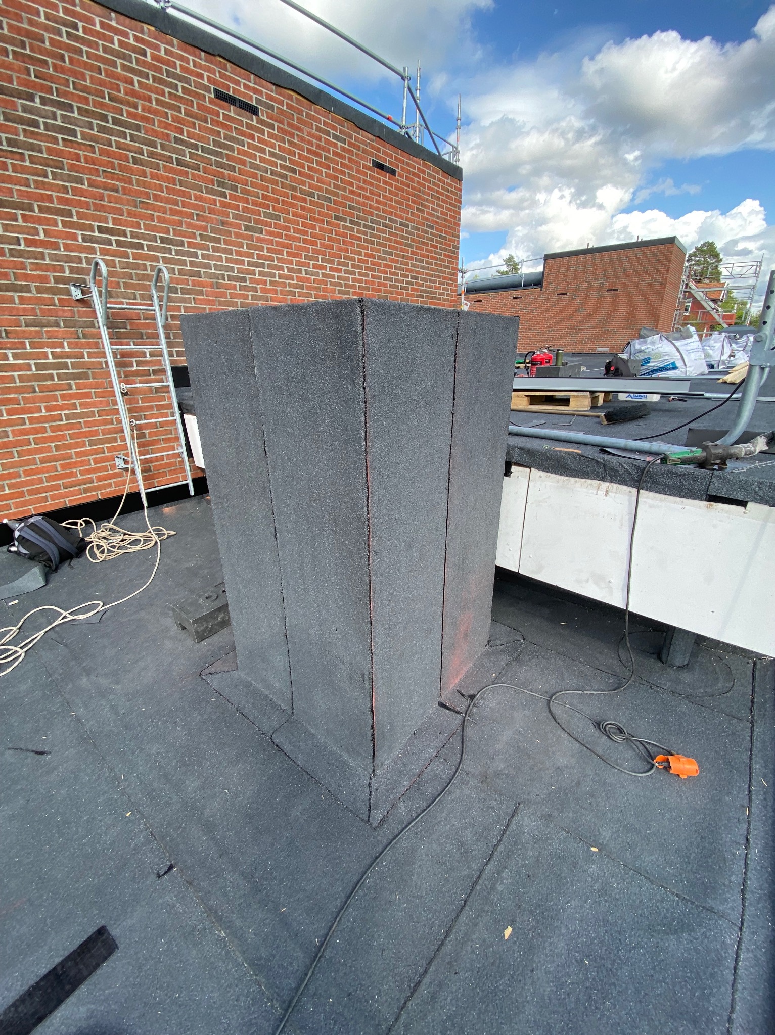 Detal z dachu krytego papą termozgrzewalną, z kominem zabezpieczonym izolacją. W tle mur z czerwonej cegły i niebo z chmurami.