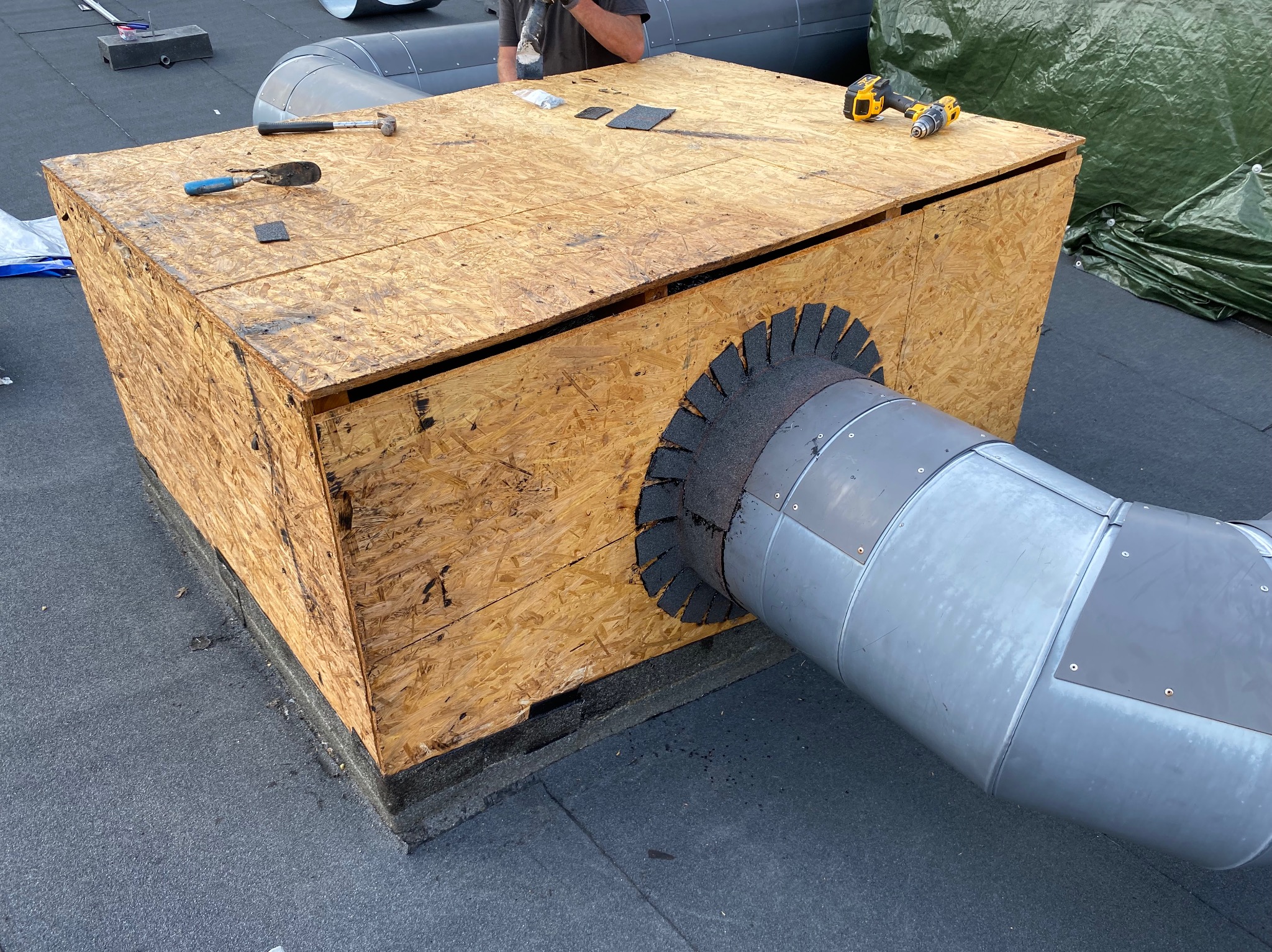 Montaż elementu wentylacyjnego na dachu płaskim, obudowanego płytą OSB, z widocznymi narzędziami i elementami uszczelniającymi. Praca w toku.