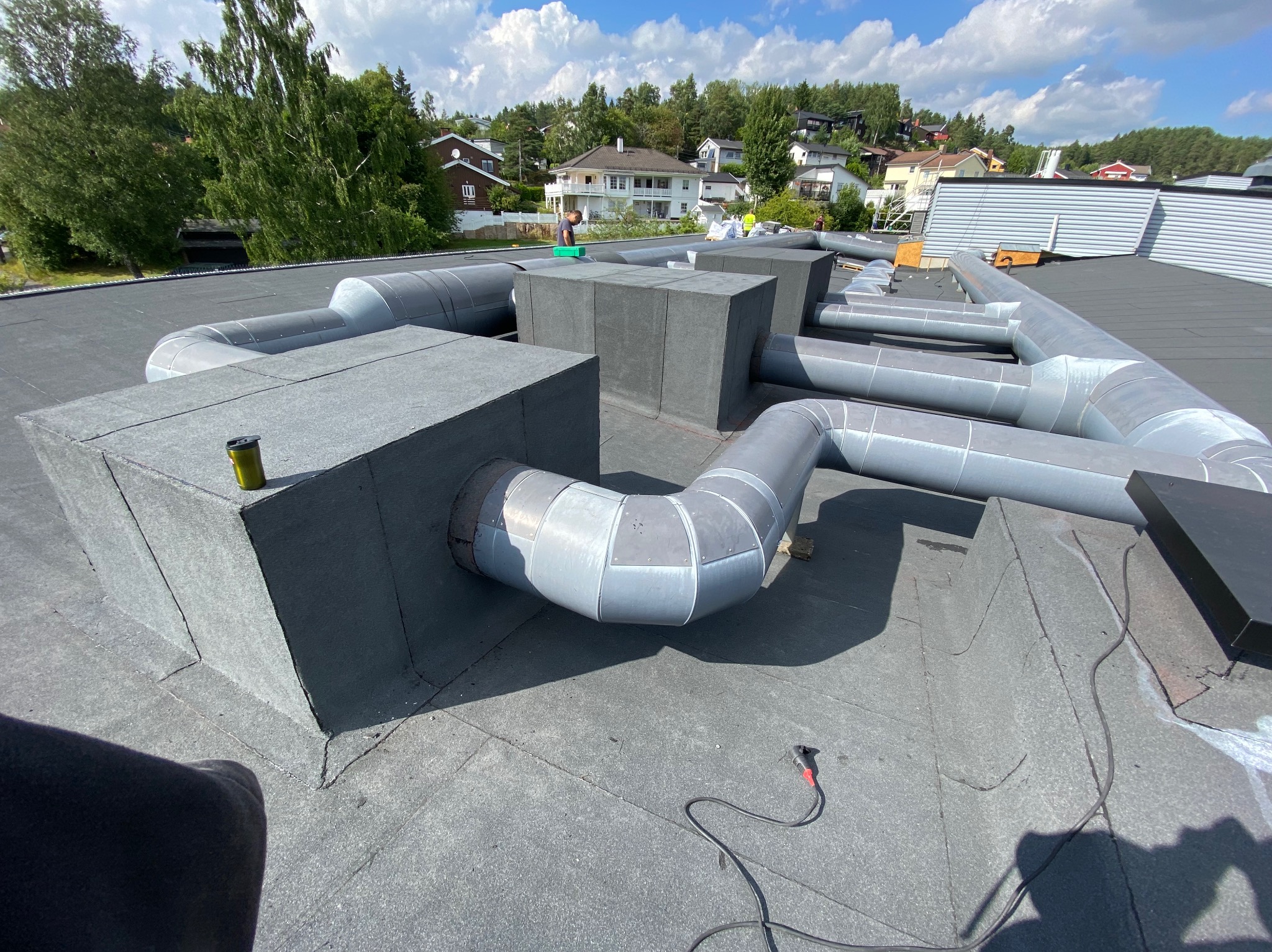 Montaż instalacji wentylacyjnej na płaskim dachu pokrytym papą. Widoczne elementy wentylacyjne, rury i narzędzia. W tle zabudowa jednorodzinna i zieleń.