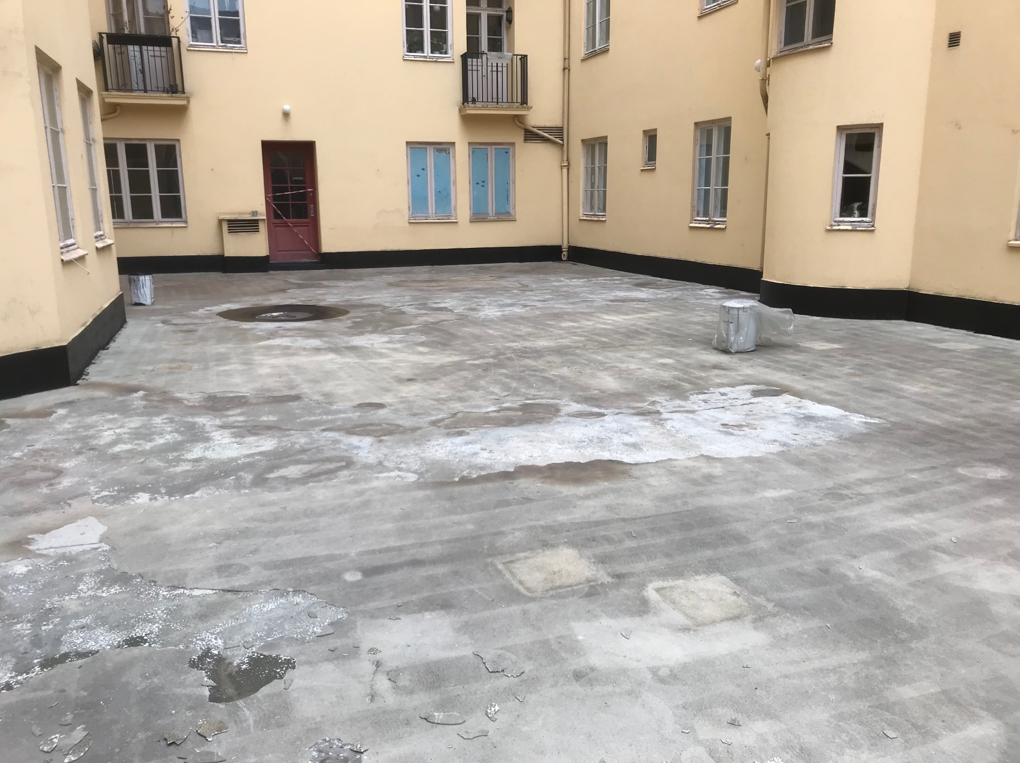 Rozległy dach płaski z widocznymi śladami wilgoci i napraw, otoczony budynkiem o żółtej elewacji z oknami i czerwonymi drzwiami. Widoczne elementy izolacji.