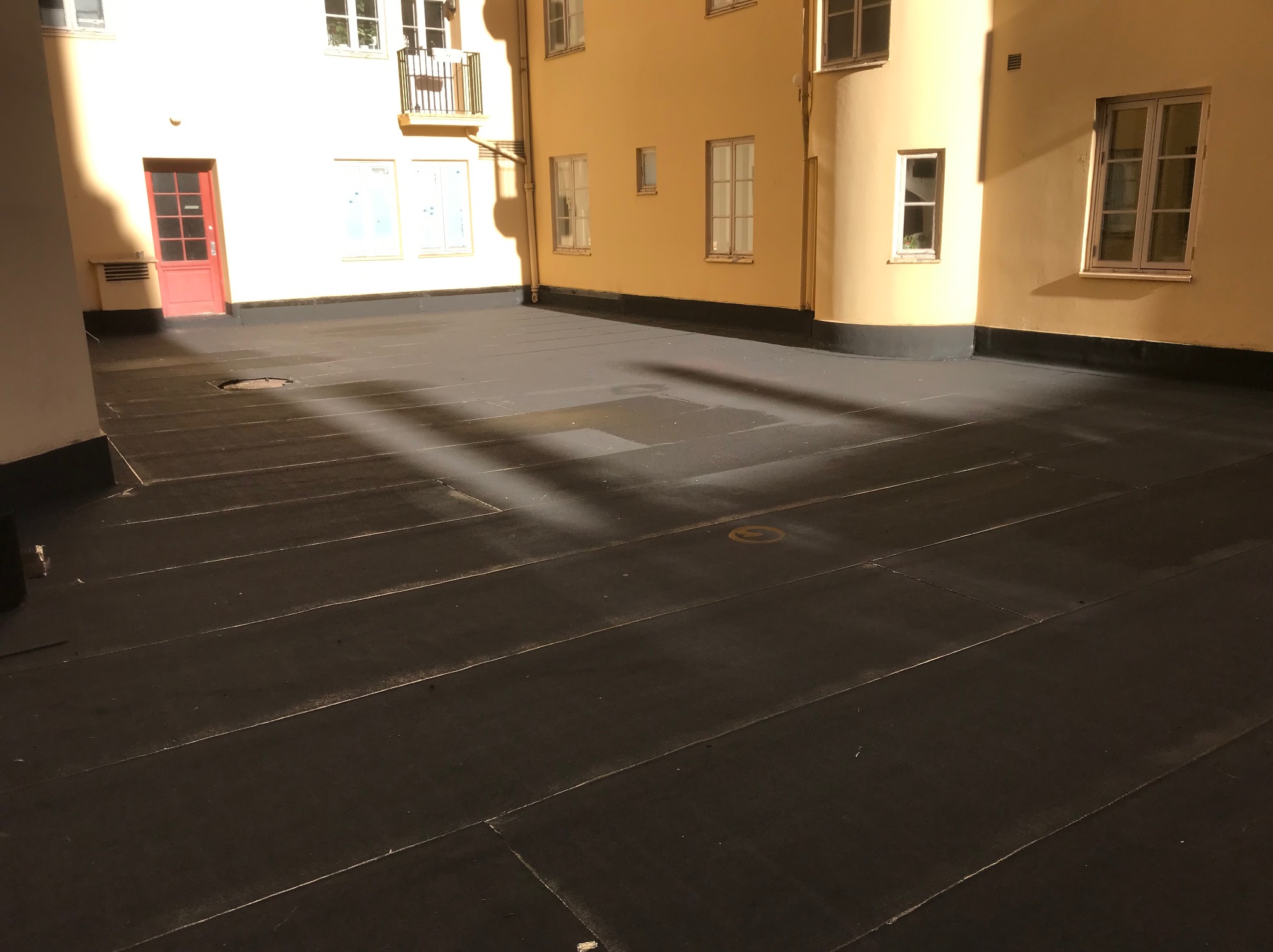 Czarny dach płaski, pokryty papą, otoczony budynkami w ciepłych odcieniach. Widoczne detale architektoniczne i gra światła na powierzchni dachu.