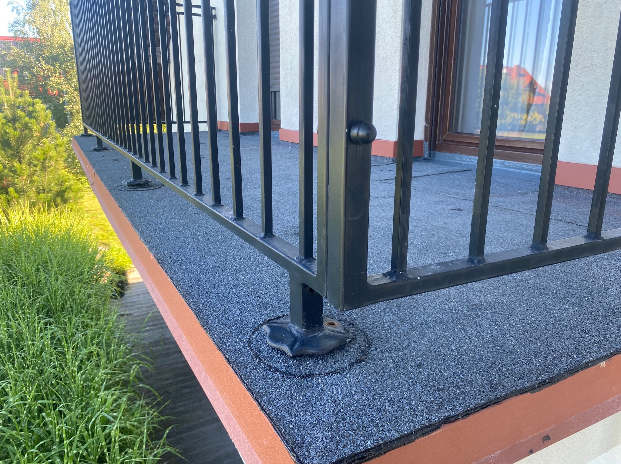 Widok z góry na fragment balkonu z czarną, metalową balustradą, pokrytego szarą papą i wykończonego brązową listwą. W tle zieleń drzew i krzewów.