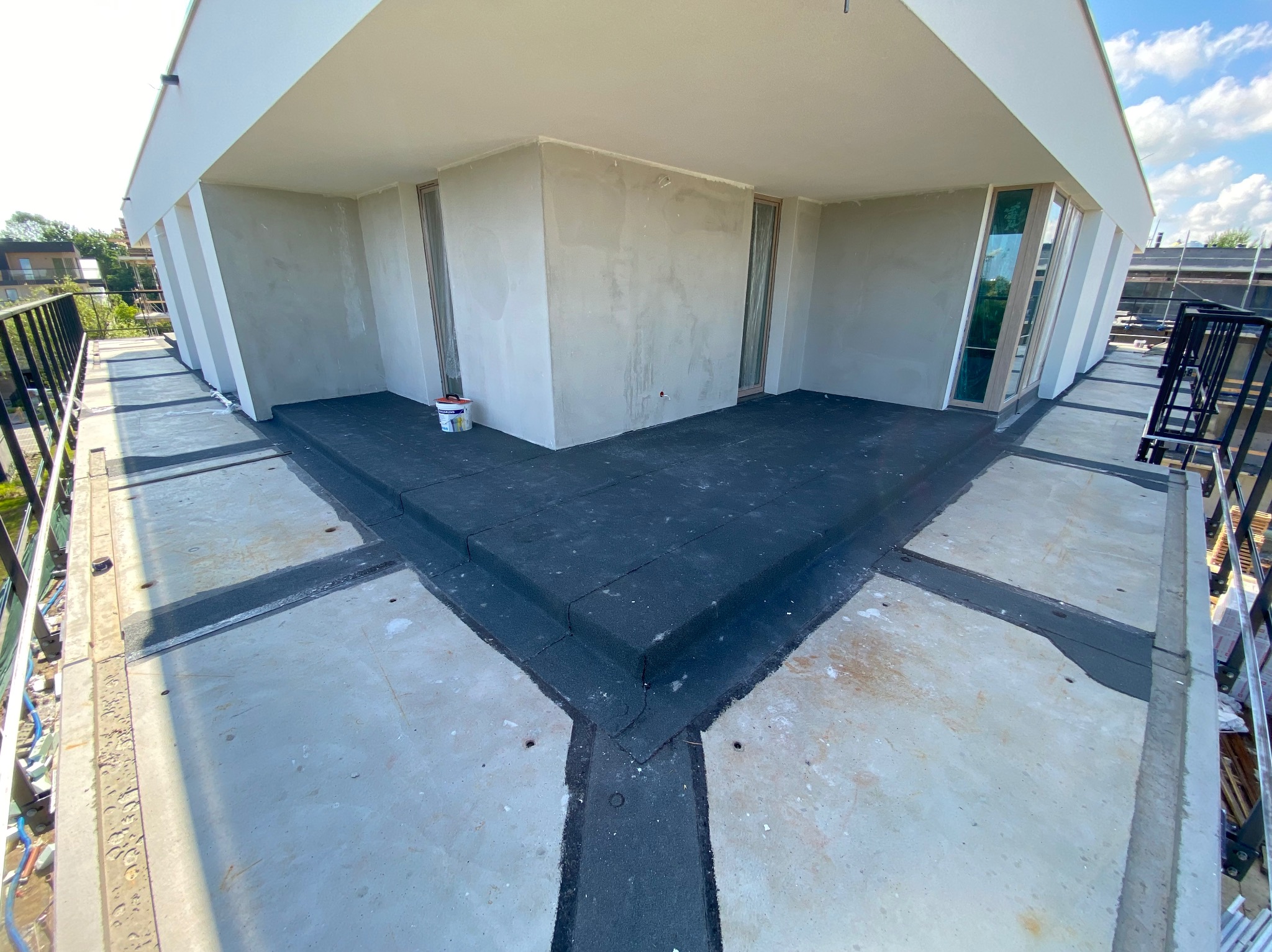 Wykończony, płaski dach z czarną izolacją, widoczne detale połączeń. Na dachu stoi puszka z farbą. Widok z góry na jasny, betonowy dach.