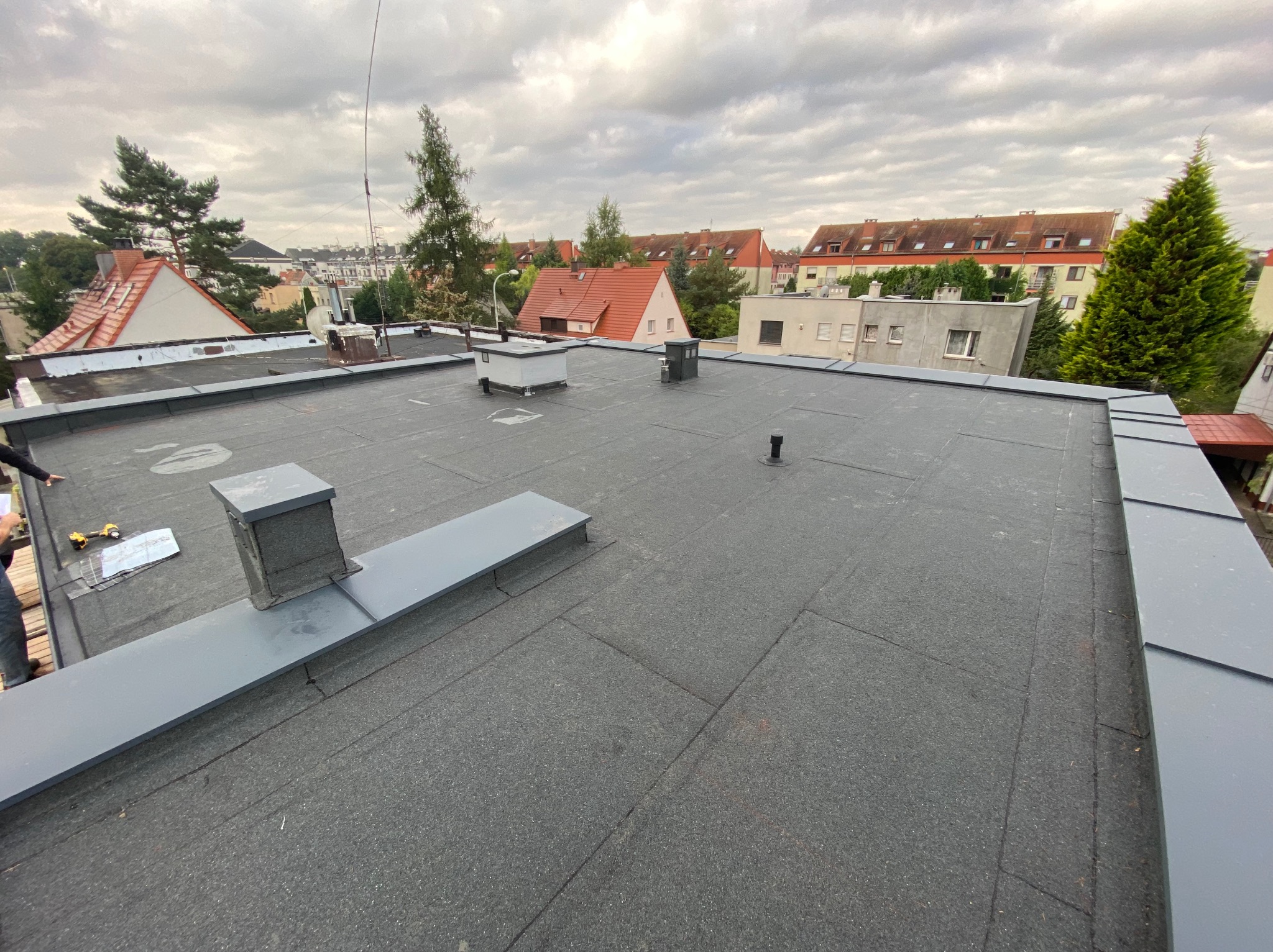 Widok z góry na płaski dach pokryty papą, z elementami obróbek blacharskich w kolorze szarym. W tle zabudowa miejska i pochmurne niebo.
