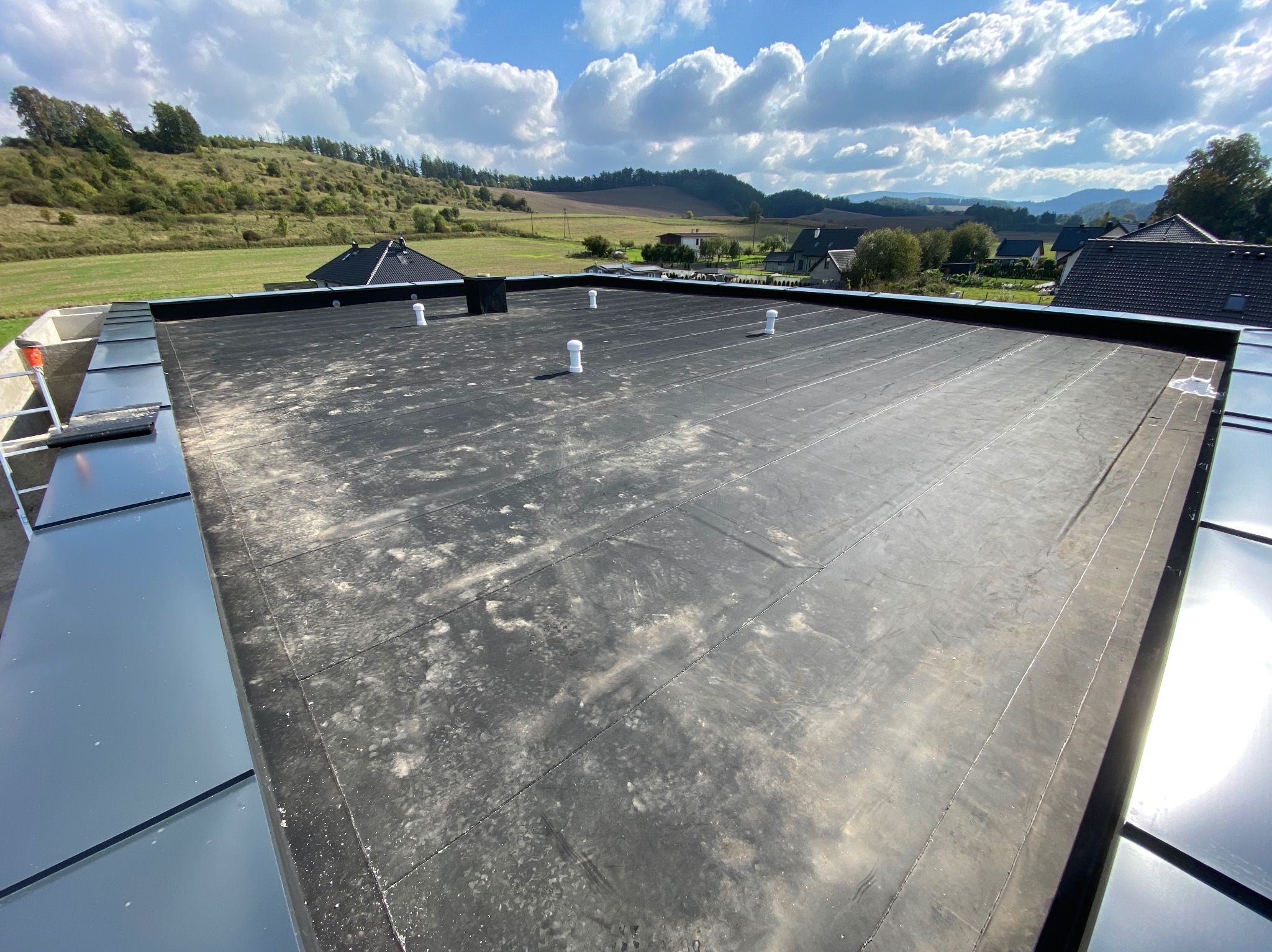 Czarny, płaski dach z papy termozgrzewalnej z kominkami wentylacyjnymi na tle zielonych wzgórz i błękitnego nieba z chmurami. Widoczne obróbki blacharskie.