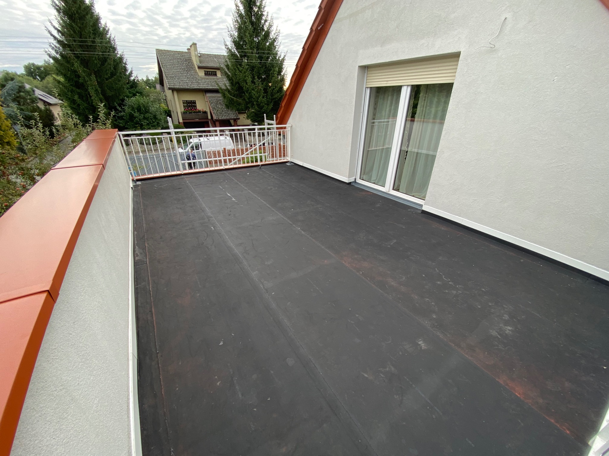 Balkon z nową, czarną hydroizolacją, zabezpieczony białą balustradą i obróbką blacharską w kolorze ceglastym. Widok na domy i drzewa.