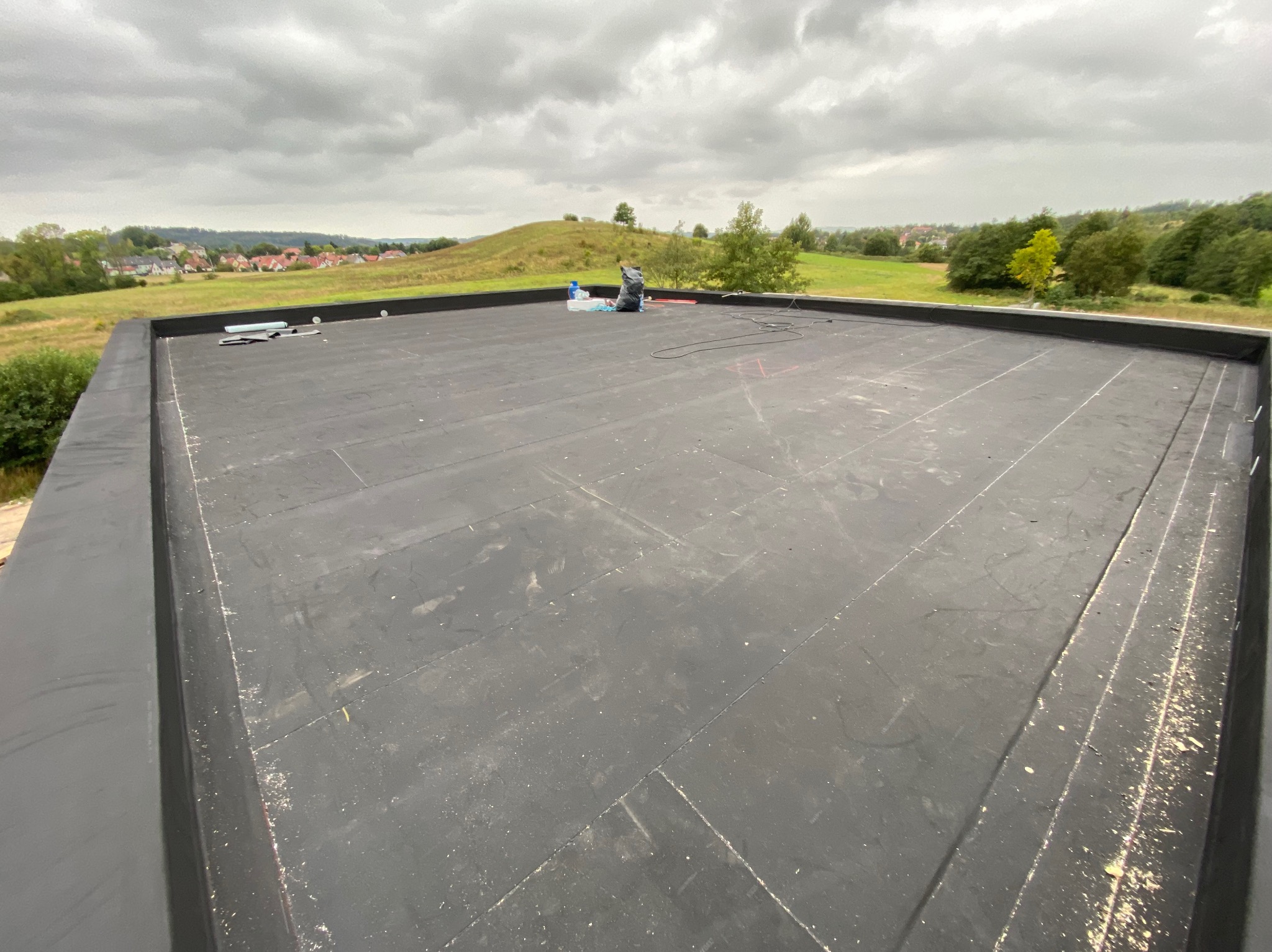 Płaski dach pokryty papą termozgrzewalną z widocznymi łączeniami. Na dachu narzędzia i materiały, w tle krajobraz z pagórkami i zabudowaniami. Pochmurne niebo.