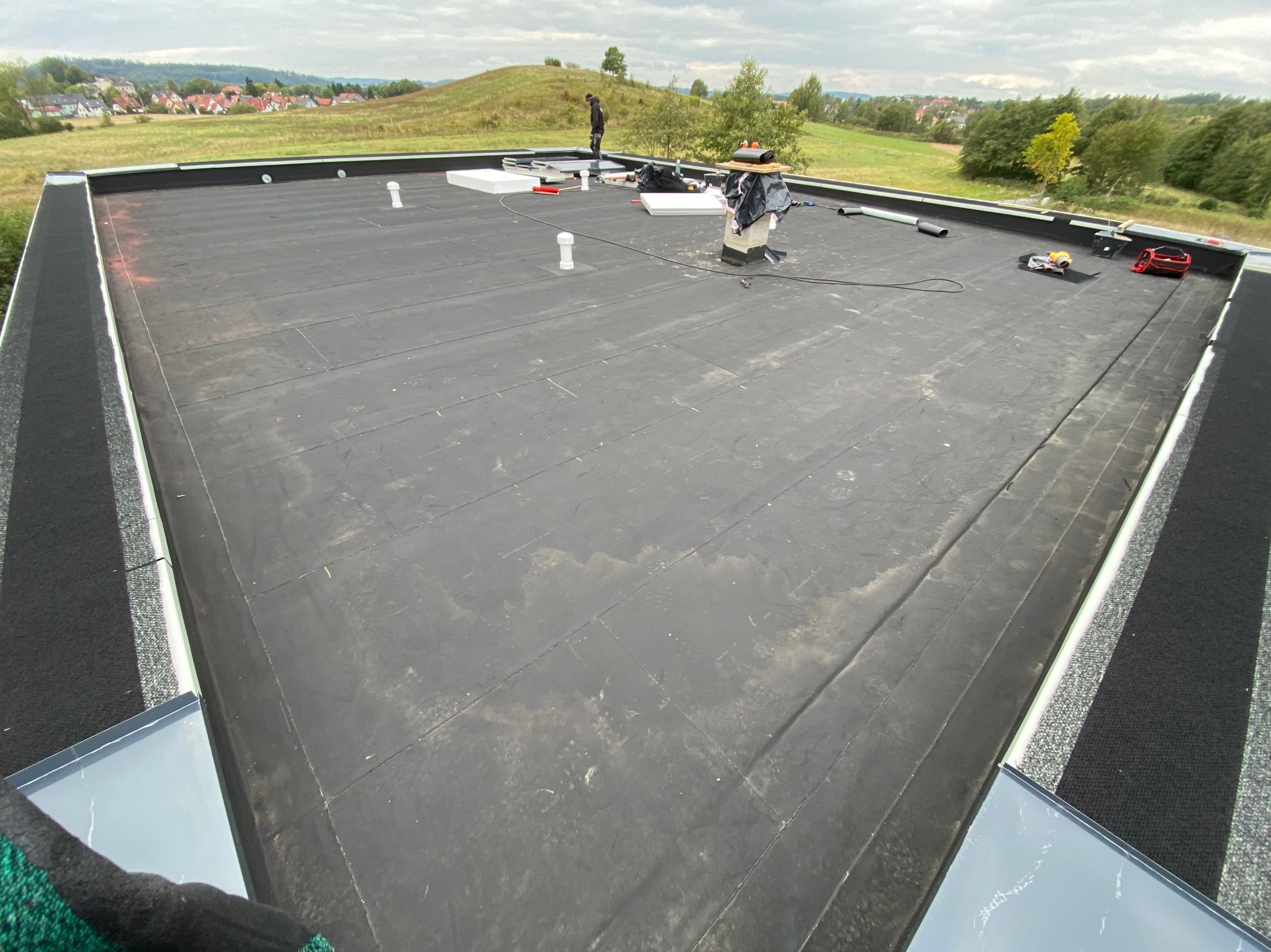 Czarny dach płaski z papy termozgrzewalnej w trakcie prac. Widoczny komin, narzędzia i sylwetka pracownika na tle zielonego krajobrazu.